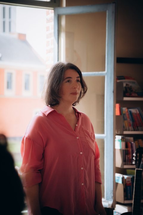 Une femme en chemise rose, debout près d’une fenêtre, dans une bibliothèque
