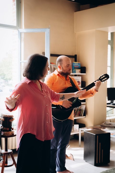 Une femme en chemise rose parle, la main tendue, à côté d’un musicien qui joue de la guitare